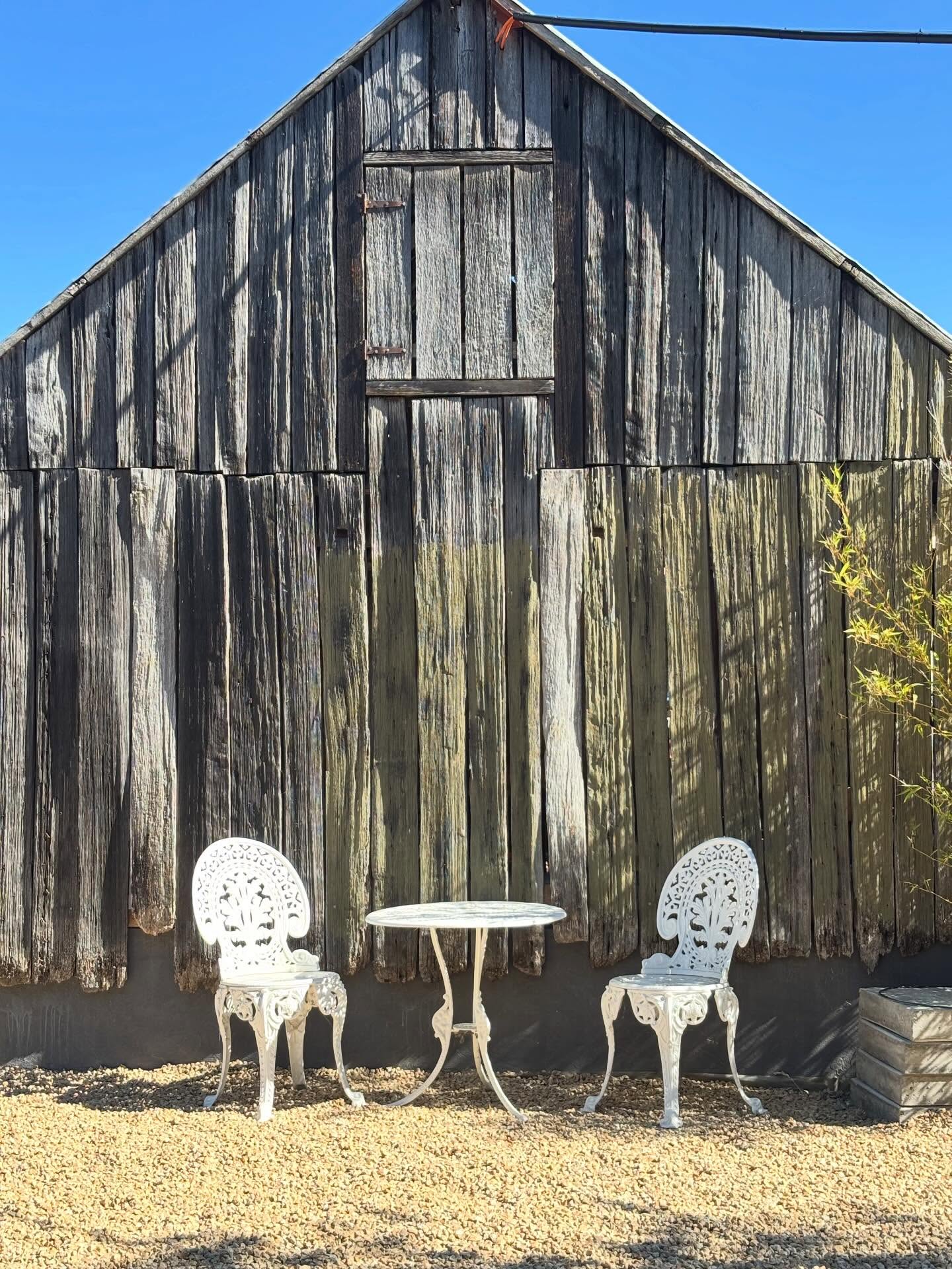 Two white wrought-iron chairs and a small table in front of a weathered timber barn